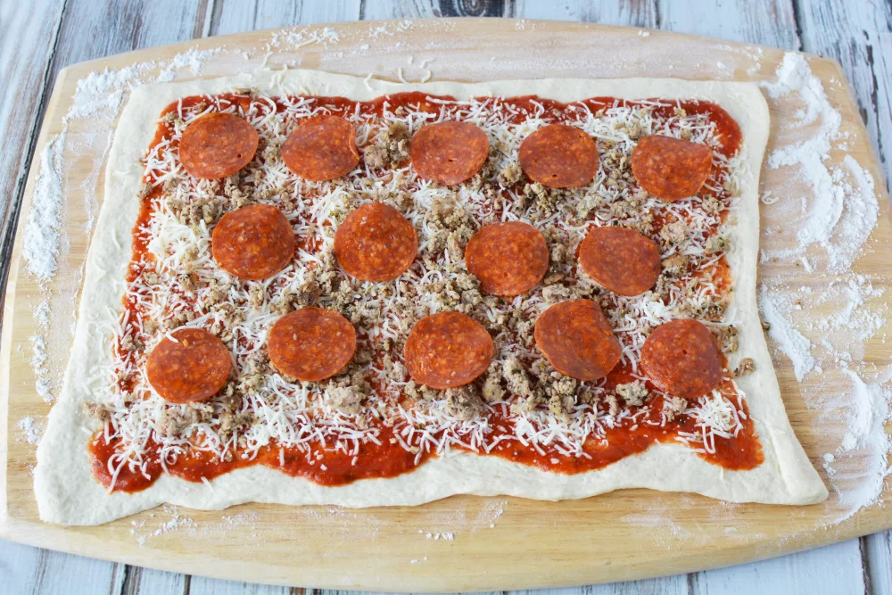 pizza sauce, cheese, pepperoni and sausage on top of a pizza dough on a cutting board.
