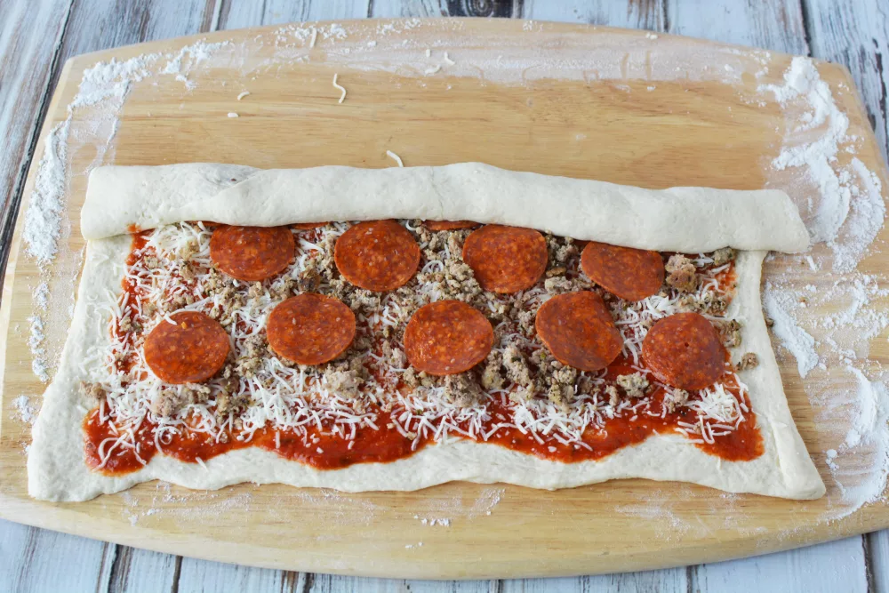 pizza sauce, cheese, pepperoni and sausage on top of a pizza dough on a cutting board being rolled up