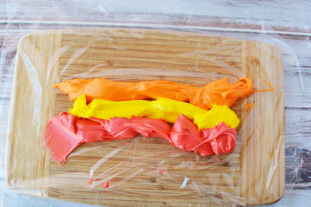 Orange, yellow, and red frosting in three rows on a sheet of plastic food wrap. 