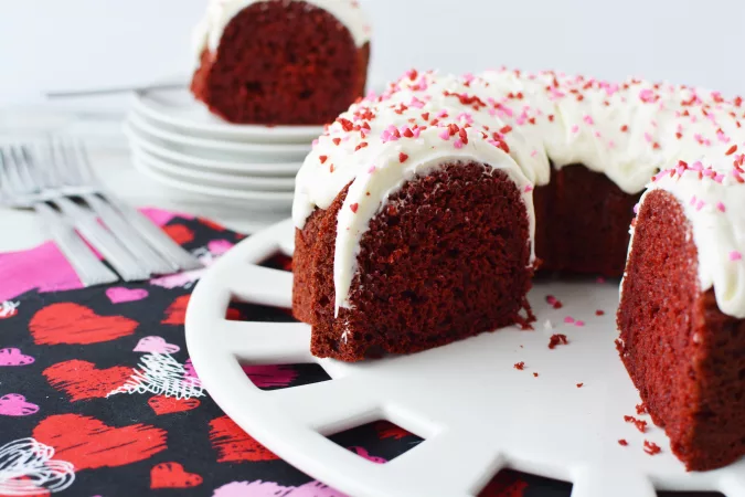 Beautiful red velvet cake with white frosting and pink and red sprinkles on a white cake plate