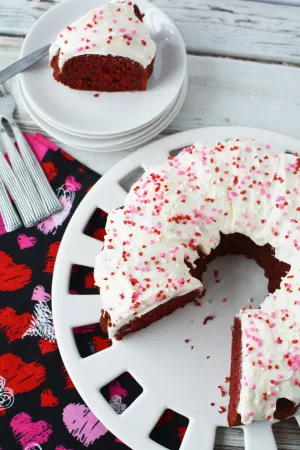 Beautiful red velvet bundt cake with white frosting and pink and red sprinkles on a white cake plate