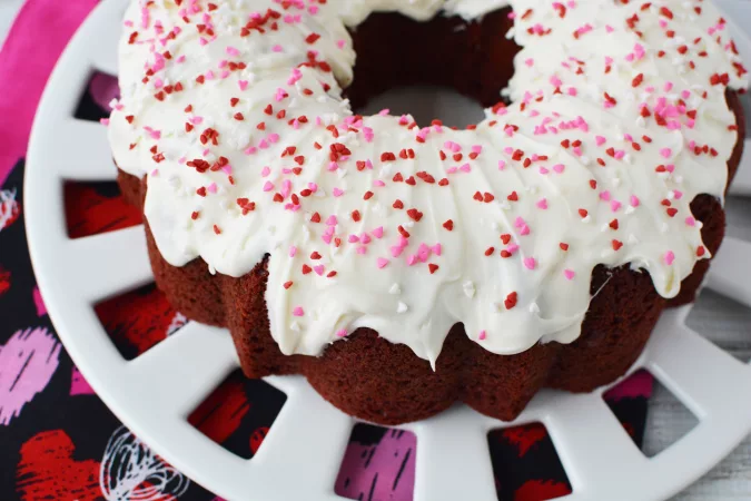Beautiful red velvet cake with white frosting and pink and red sprinkles on a white cake plate