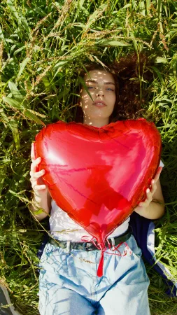 Woman holding a big red puffy heart balloon laying in a field of grass