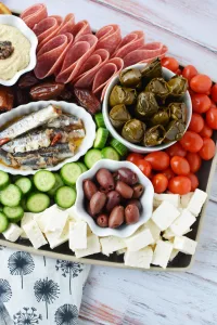 Greek Charcuterie Board Ideas a charcuterie board filled with tomatoes, cheese, meats, salami, anchovies, wrapped dates and grapes and olives