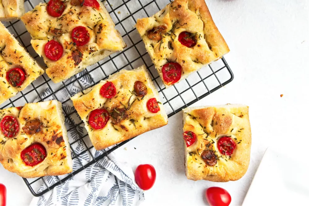 Focaccia bread topped with roasted garlic, roasted tomatoes and rosemary on a black cooling rack