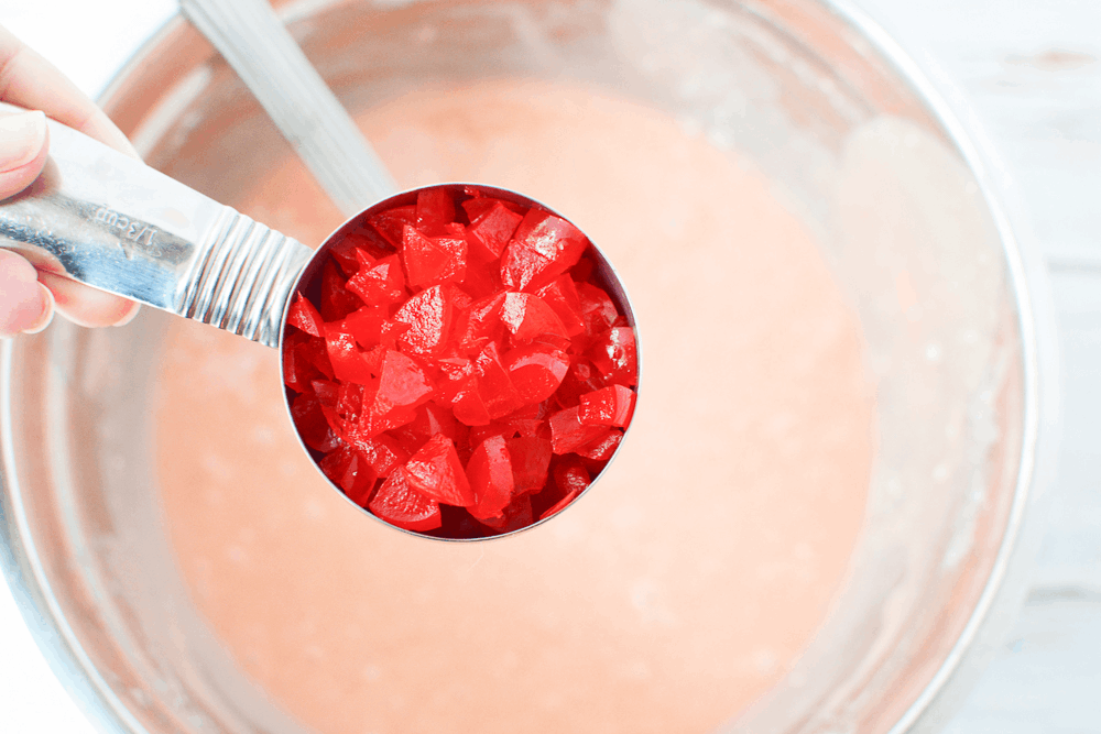 measuring spoon full of maraschino cherries being added to a bowl of cake batter