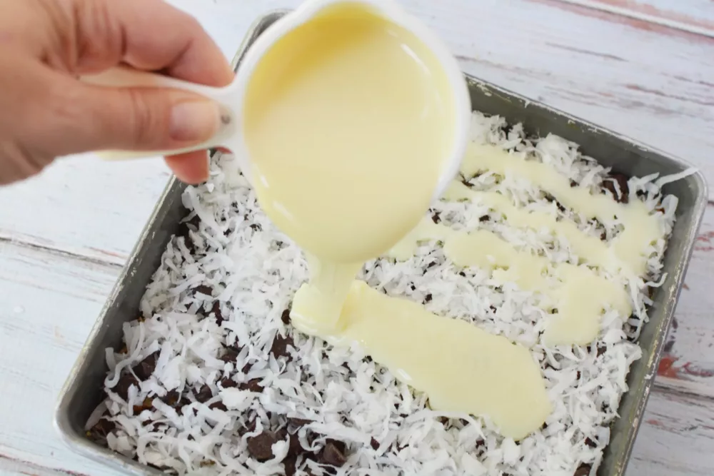 Sweetned condensed milk being poured over layered dream bar ingredients in baking dish.