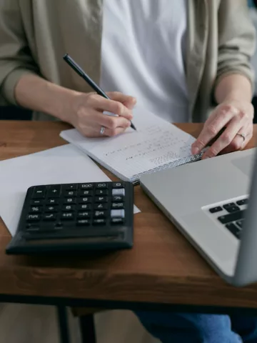 person sitting at a desk writing on a notepad with a laptop and calculator in front of them