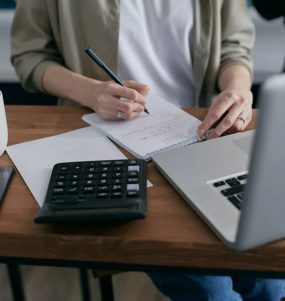 person sitting at a desk writing on a notepad with a laptop and calculator in front of them