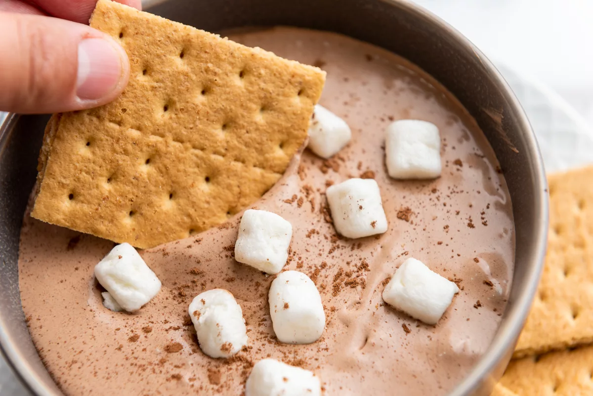 hand dipping graham cracker into hot cocoa dip with marshmallows