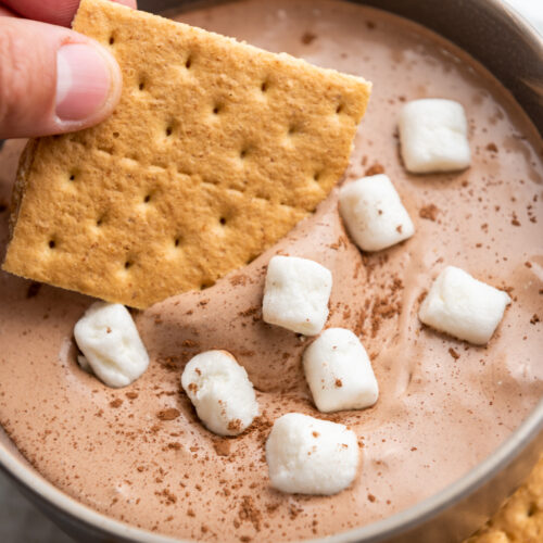 hand dipping graham cracker into hot cocoa dip with marshmallows
