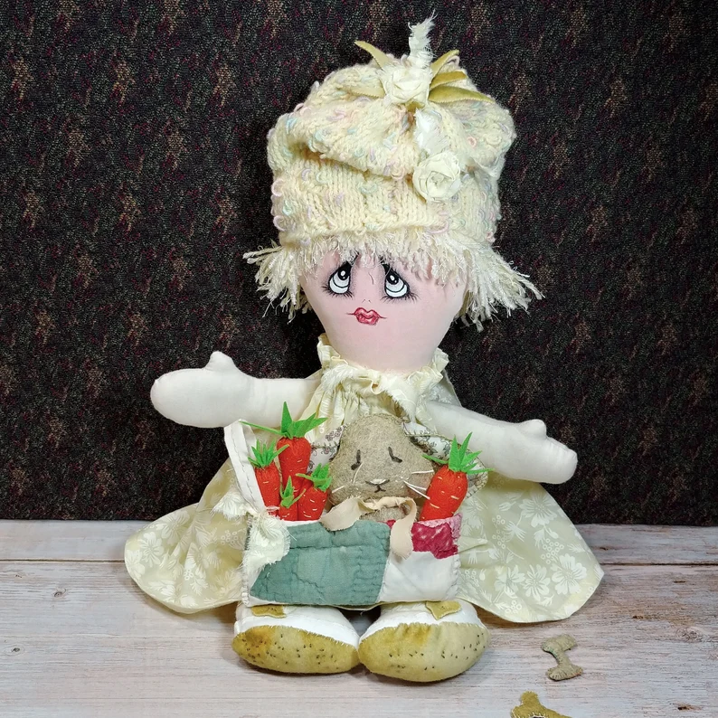 Fabric art doll with knit hat and basket with baby bunny and hand-sewn carrots. 