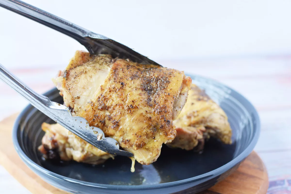 Closeup shot of tongs holding a cooked chicken thigh in front of plate with more Dutch oven chicken thighs. 
