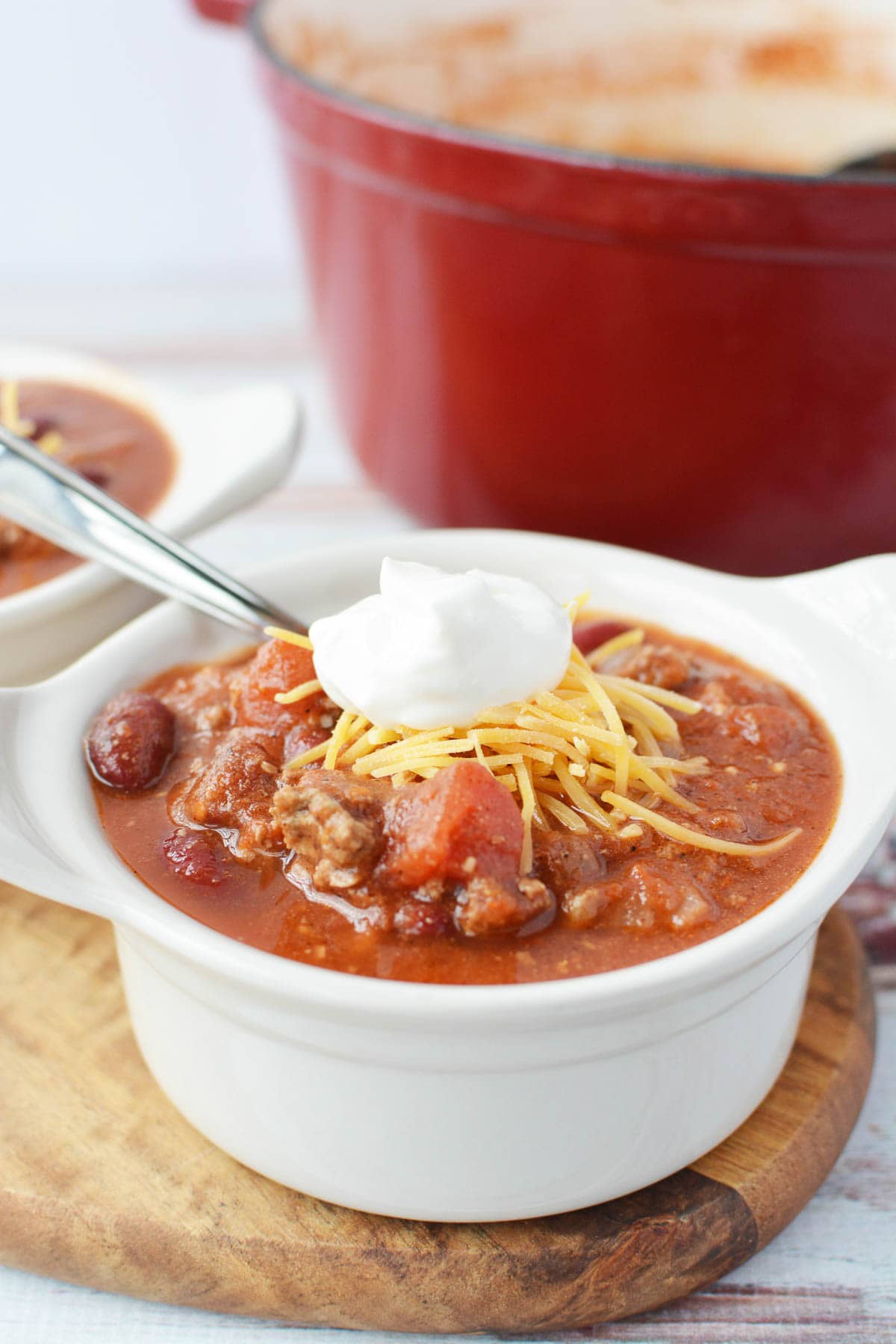 Closeup shot of bowlful of Dutch oven chili topped with cheese and sour cream with Dutch oven filled with more chili in background. 