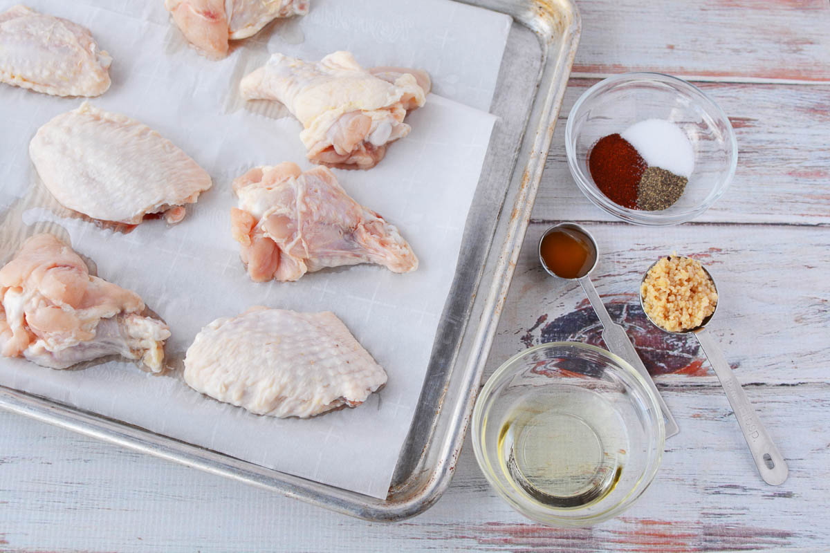 Overhed shot of baking sheet with chicken wings next to individual seasonings and oil in bowls. 