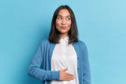 woman wearing a white shirt and blue cardigan touching her stomach while thinking about Gut Health and Probiotic Drinks