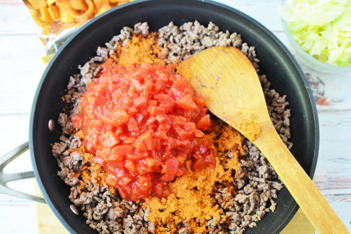 skillet filled with cooked ground beef and topped with taco seasoning and diced tomatoes with a wooden spoon